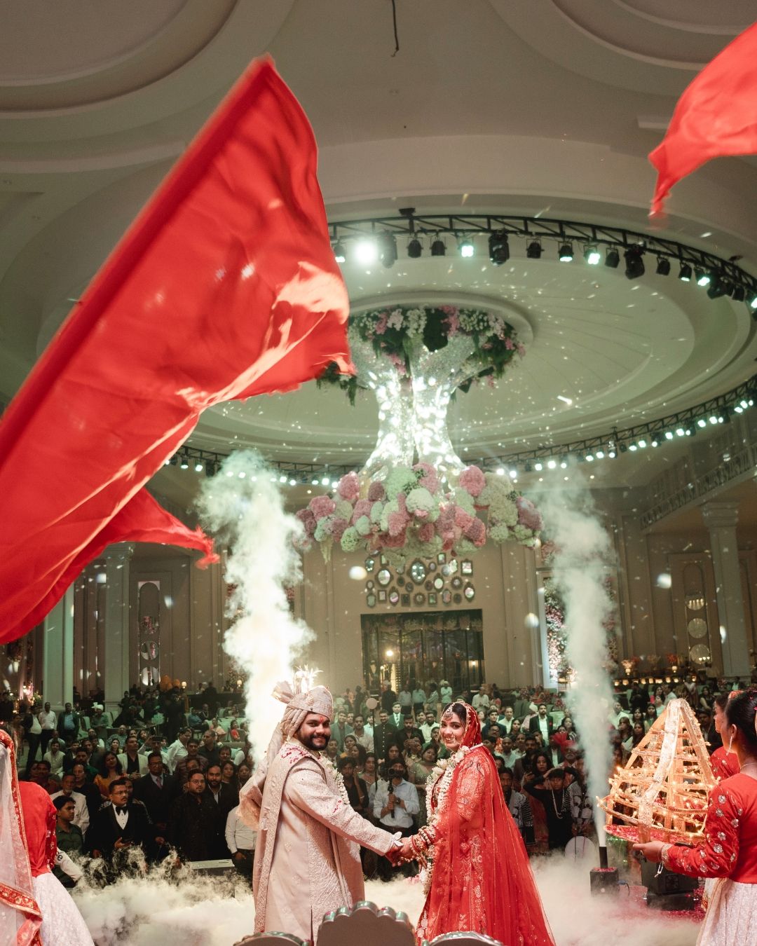 Indian bride and groom in red attire hold hands at grand wedding ceremony with smoke effects and floral chandelier.