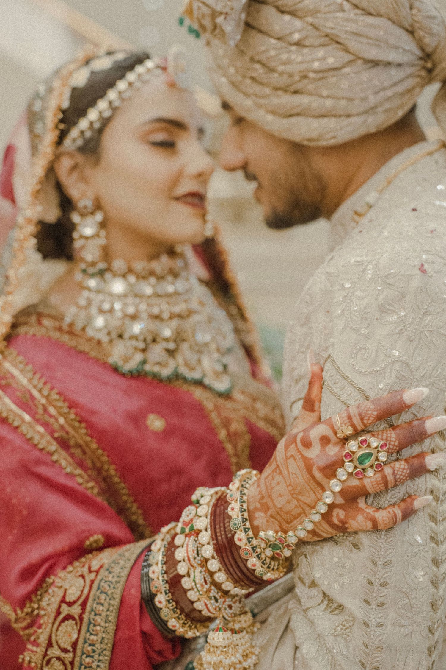 Indian bride and groom in traditional wedding attire, bride wearing red lehenga with gold jewelry and mehndi hands.