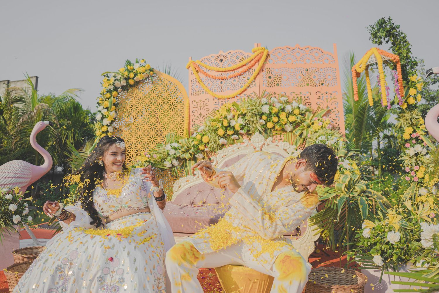 Indian couple in yellow and white traditional attire celebrating Haldi ceremony with floral decor and flamingo backdrop.