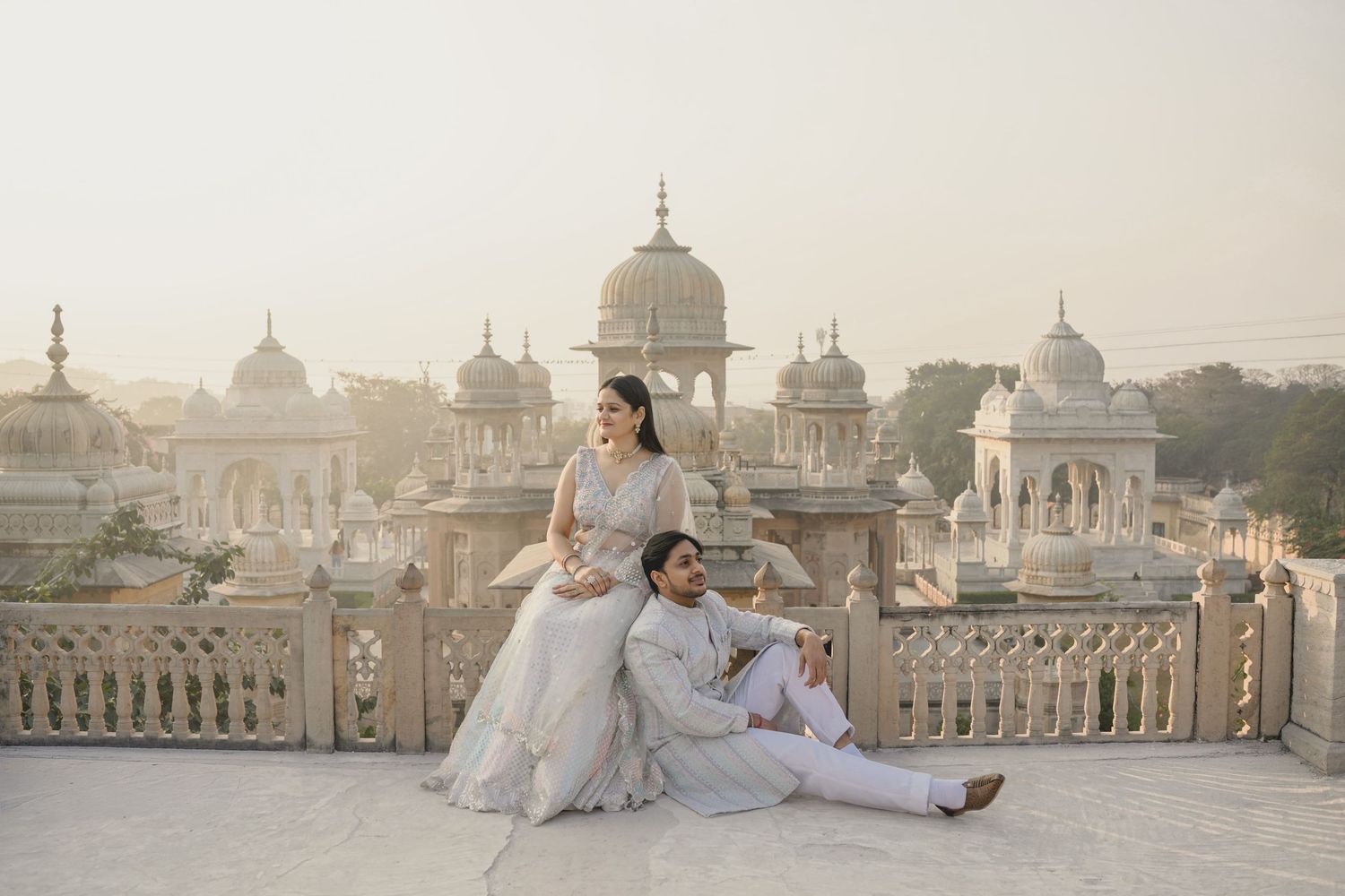 Couple in white traditional Indian attire posing on a rooftop terrace with ornate marble architecture in Udaipur.