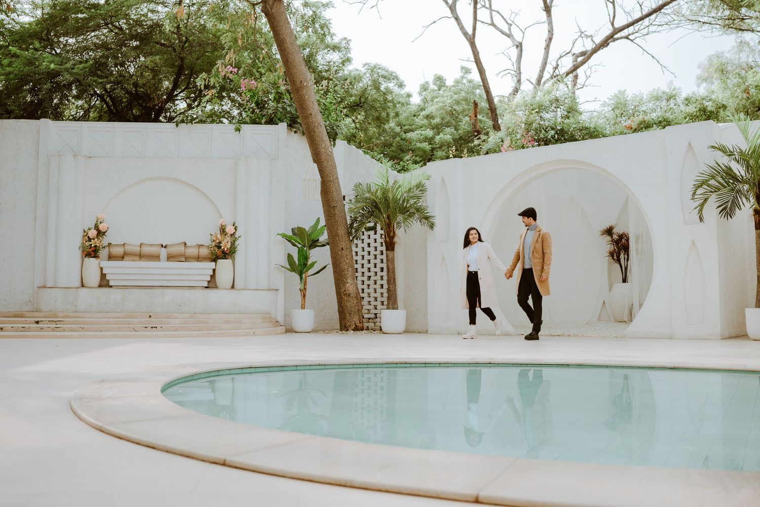 Couple walking hand in hand beside a white-walled pool area with arched alcoves and tropical plants.
