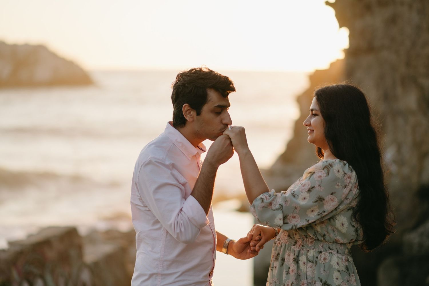 A man kisses a woman's hand at sunset on a rocky beach in a romantic engagement photo.