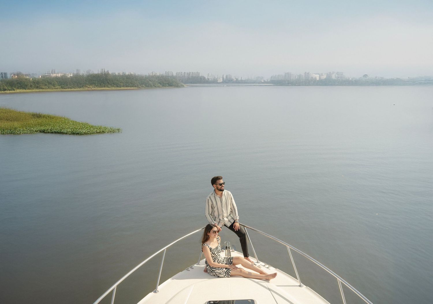 Couple relaxing on the bow of a luxury yacht on a calm lake with a green island and city skyline in the background.
