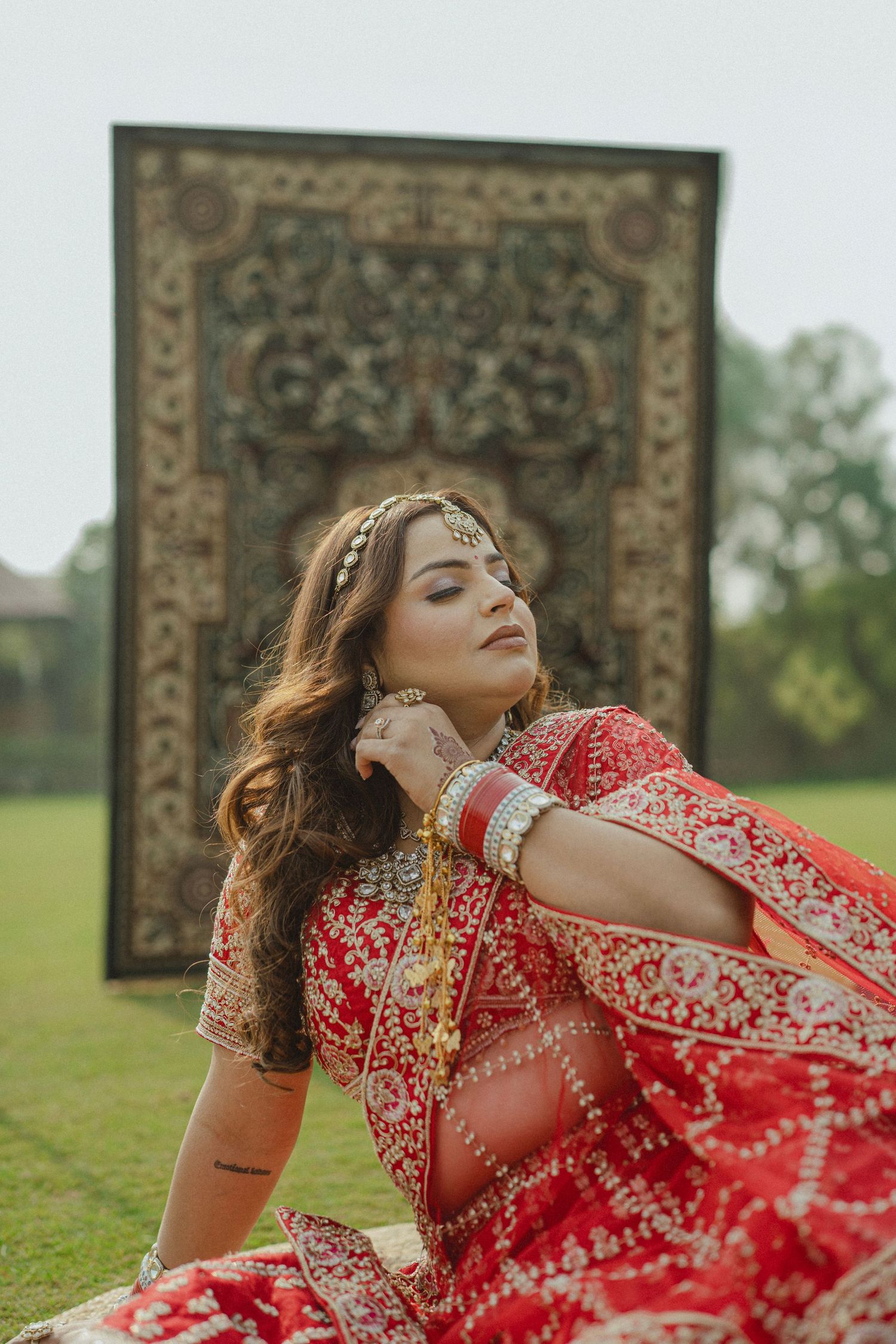 Bride in red embroidered lehenga with gold jewelry poses outdoors before ornate tapestry backdrop.