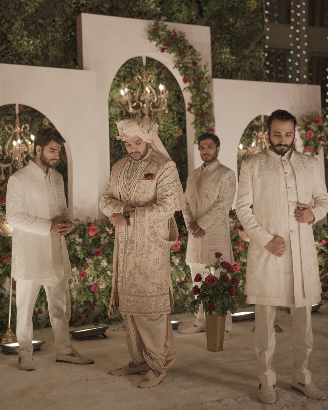 Four men in elegant cream sherwanis pose at an Indian wedding celebration with floral arch backdrop.