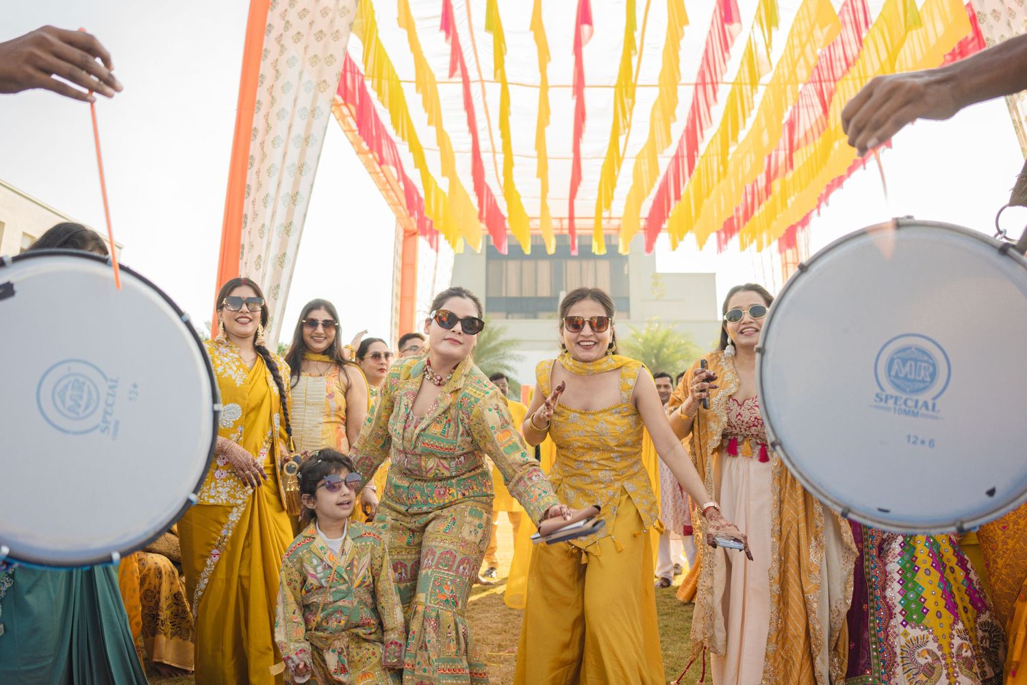 Women in yellow outfits dancing at a vibrant Indian wedding celebration under colorful floral decorations.