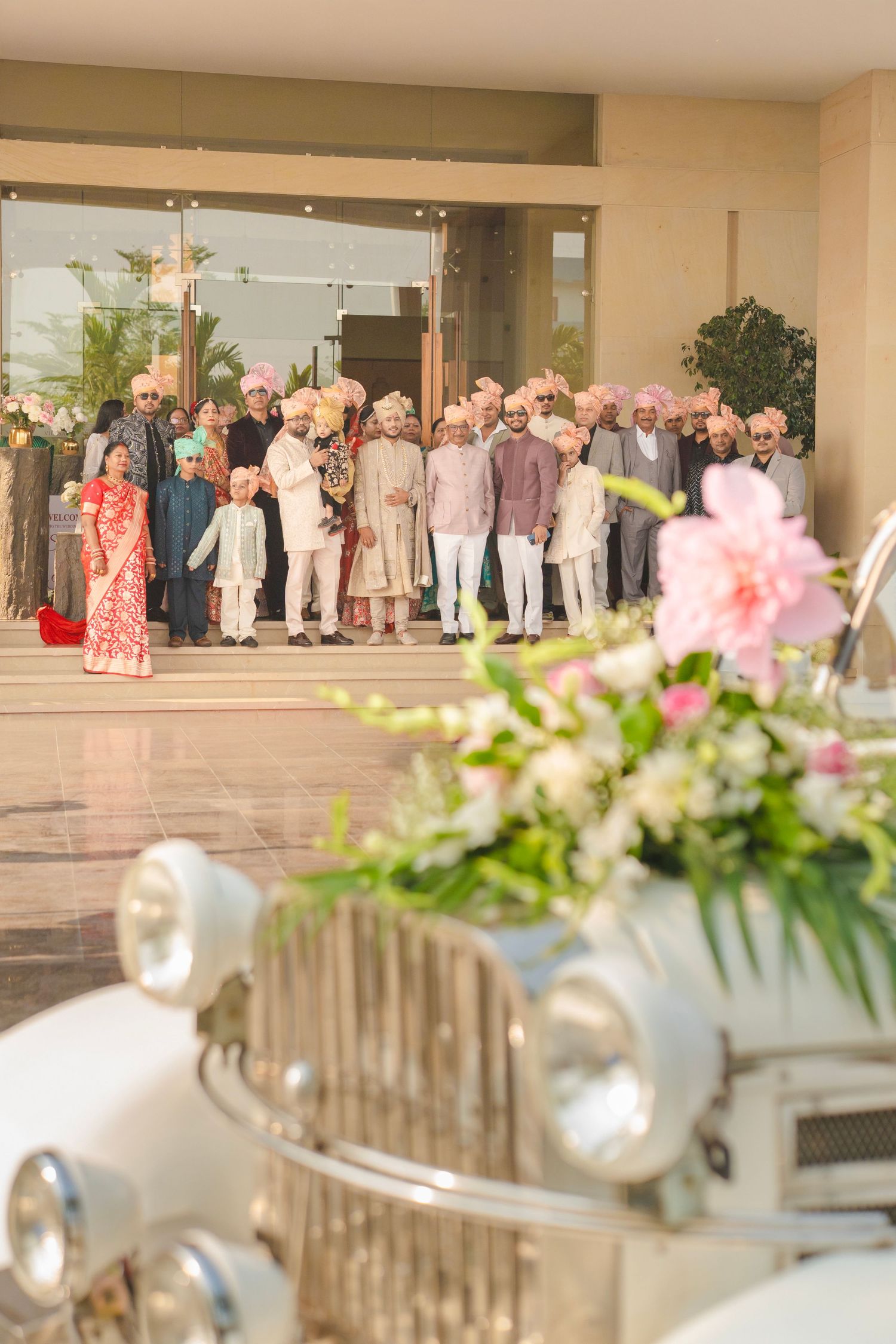 Indian wedding party in traditional attire poses outside venue, with vintage white car and floral arrangement in foreground.
