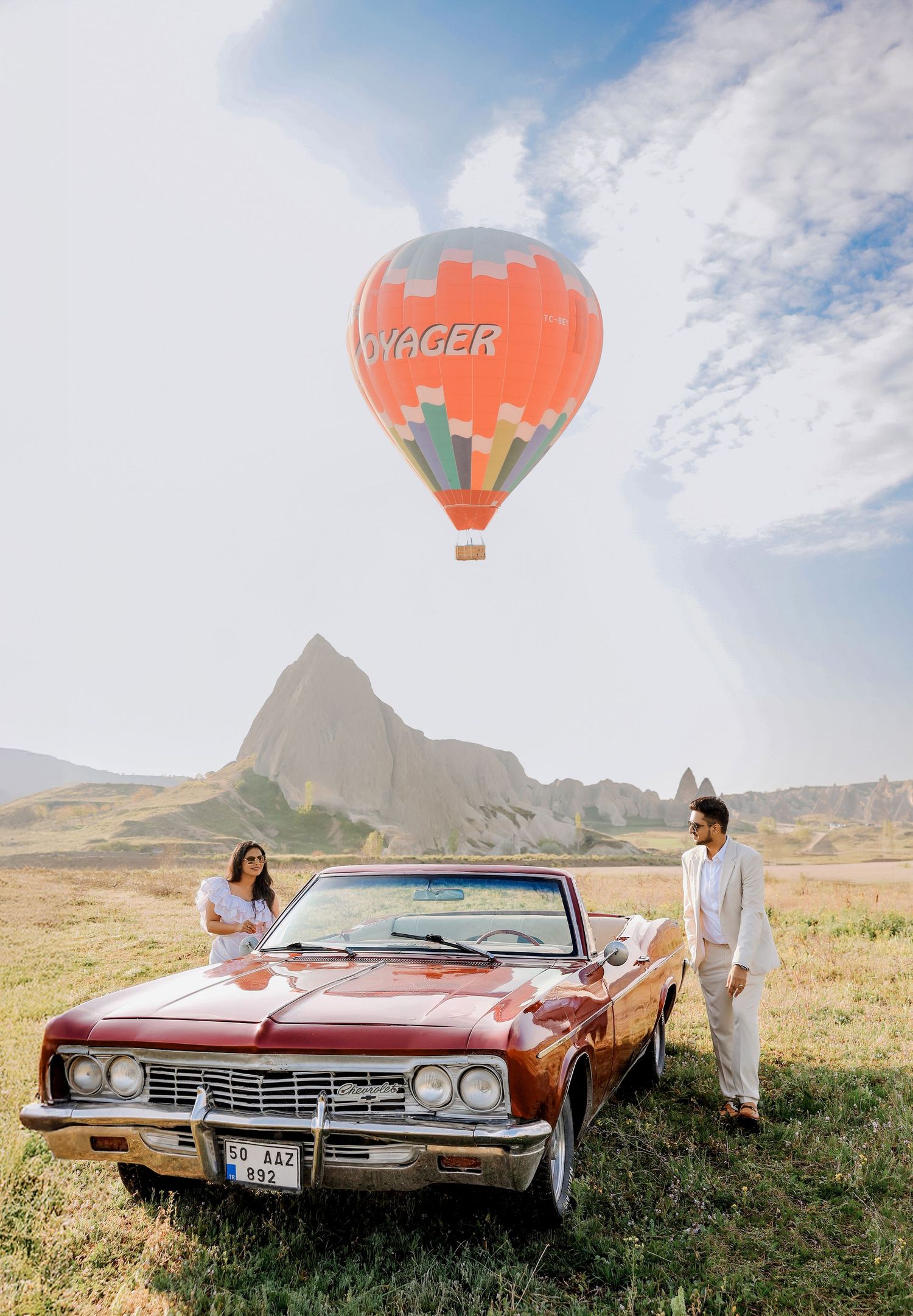 Vintage red car parked in a field with hot air balloons floating in the blue sky.