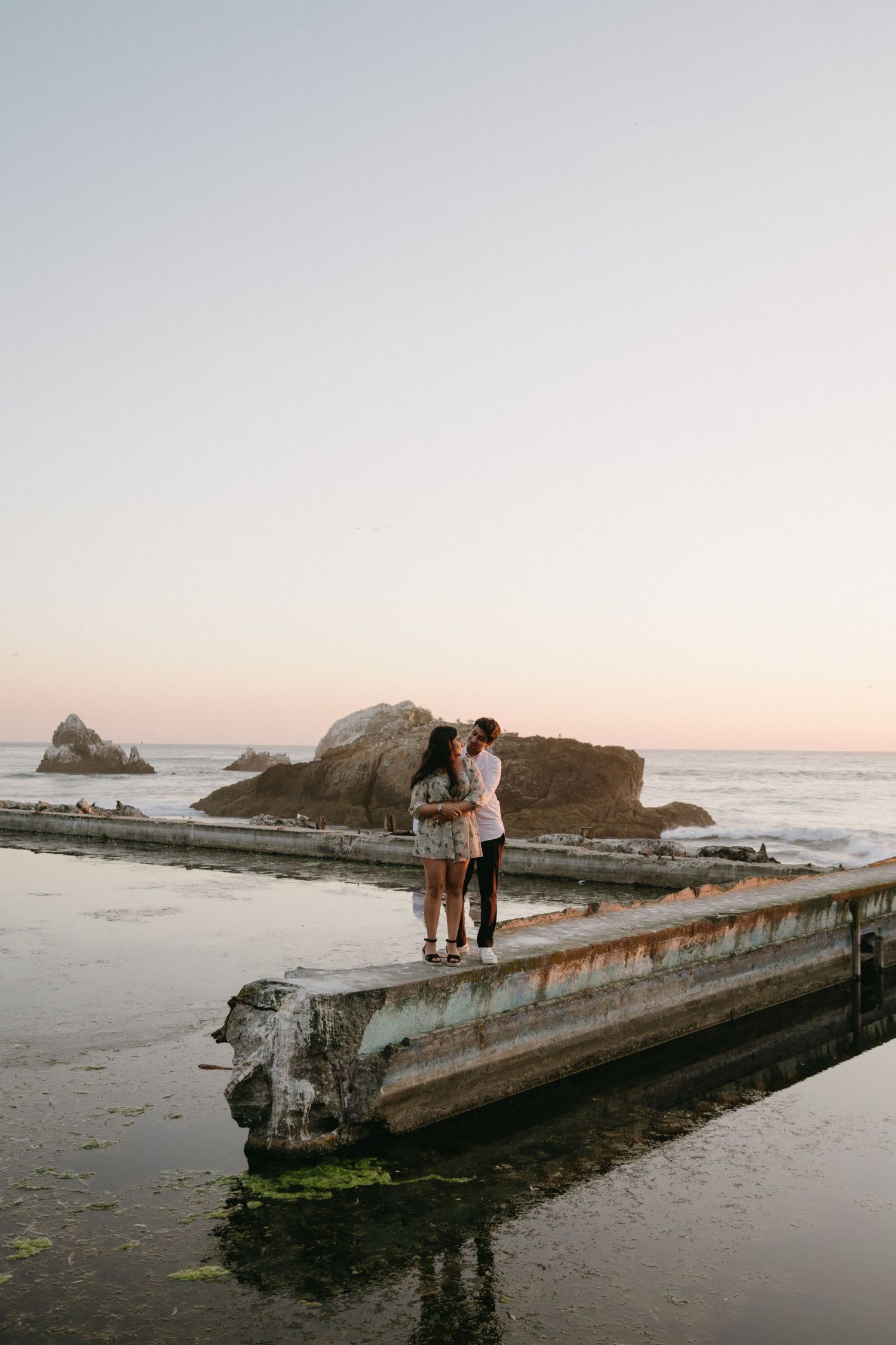 Couple embracing at sunset on a coastal concrete pier with rocky ocean backdrop at dusk.