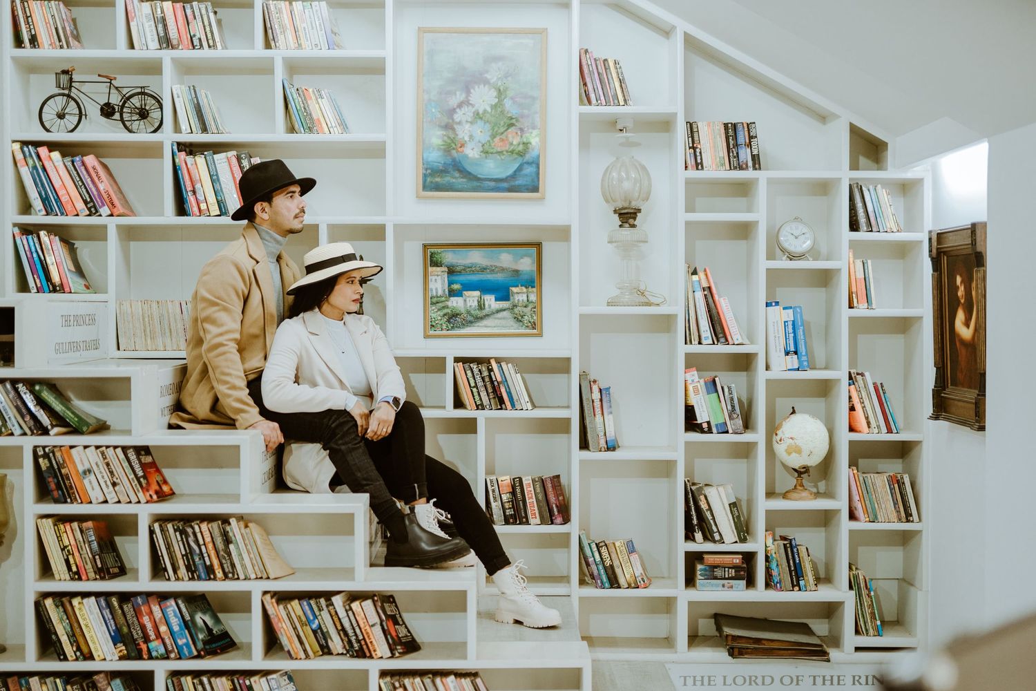 Two people sitting on white built-in bookshelves filled with colorful books in a cozy library setting.