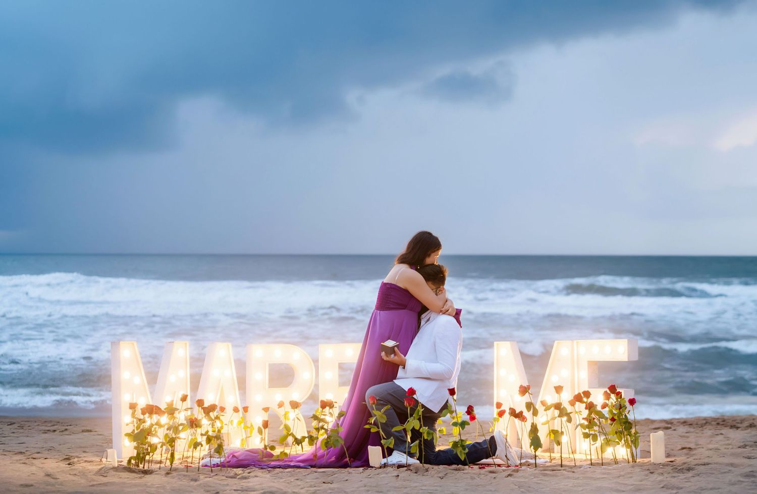 Beach proposal setup at sunset with illuminated letters and candles along the shoreline.
