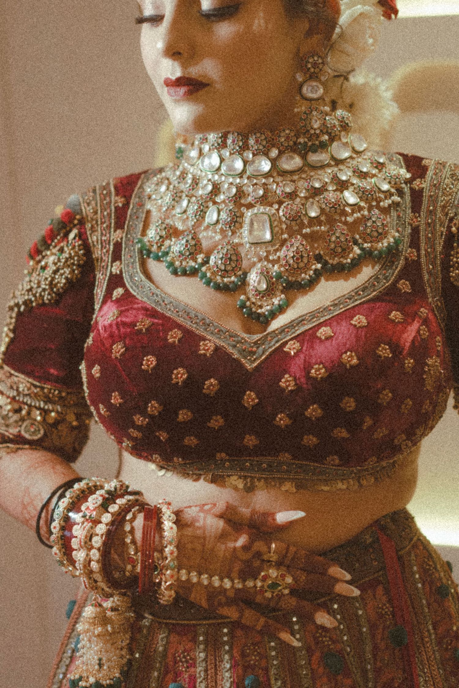 Indian bride in red lehenga with gold embroidery, layered kundan necklace, bangles, and floral hair accessories.