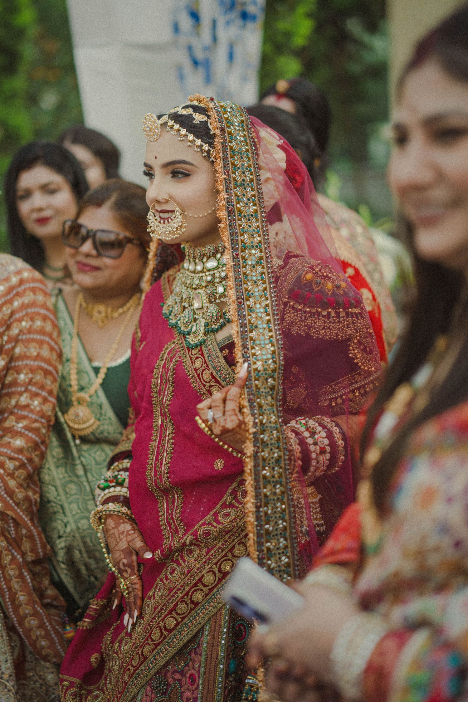 Bride in red bridal lehenga with gold jewelry and maang tikka walks with guests at a South Asian wedding.