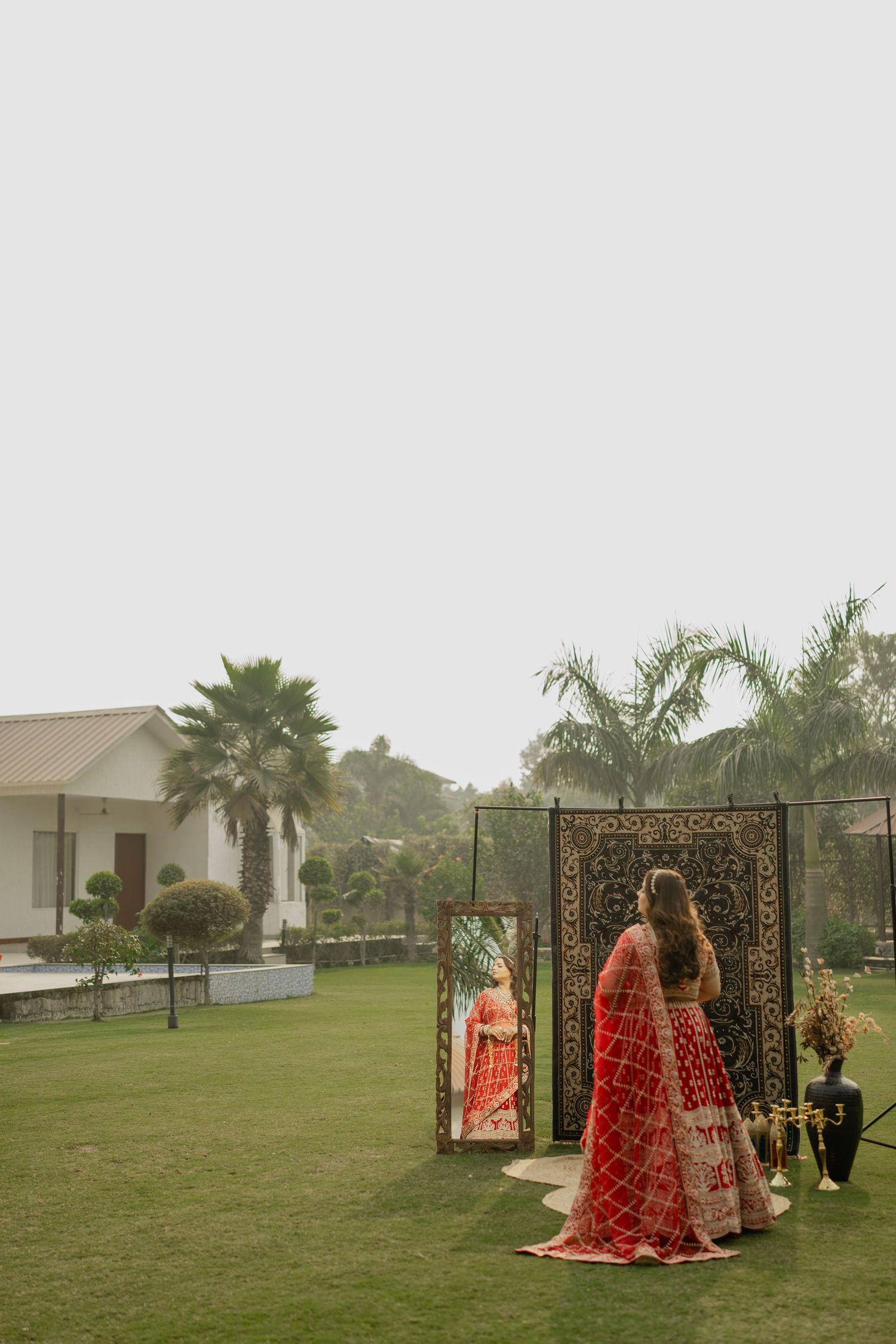 Bride in red lehenga admiring herself in ornate mirror on lush green lawn with palm trees at Indian wedding.