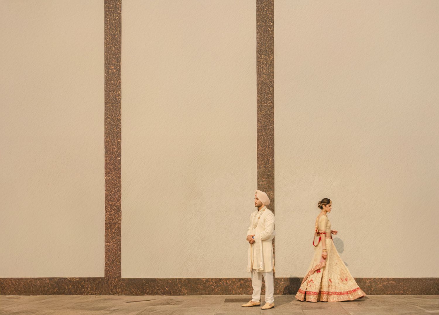 Indian bride and groom in traditional wedding attire stand apart against a minimalist beige wall.
