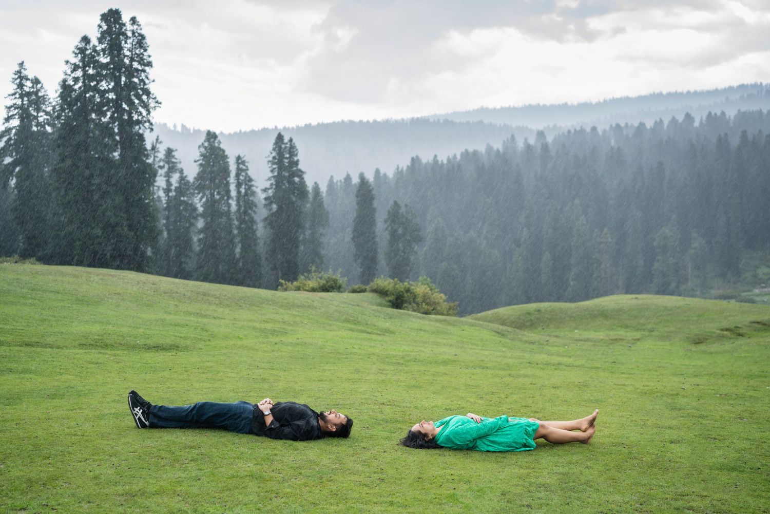 Two people lying on a lush green meadow with misty pine forest mountains in the background.