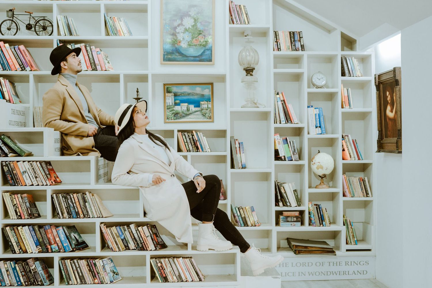 Couple sitting on white bookshelves filled with books in a cozy library setting, surrounded by artwork and decor.