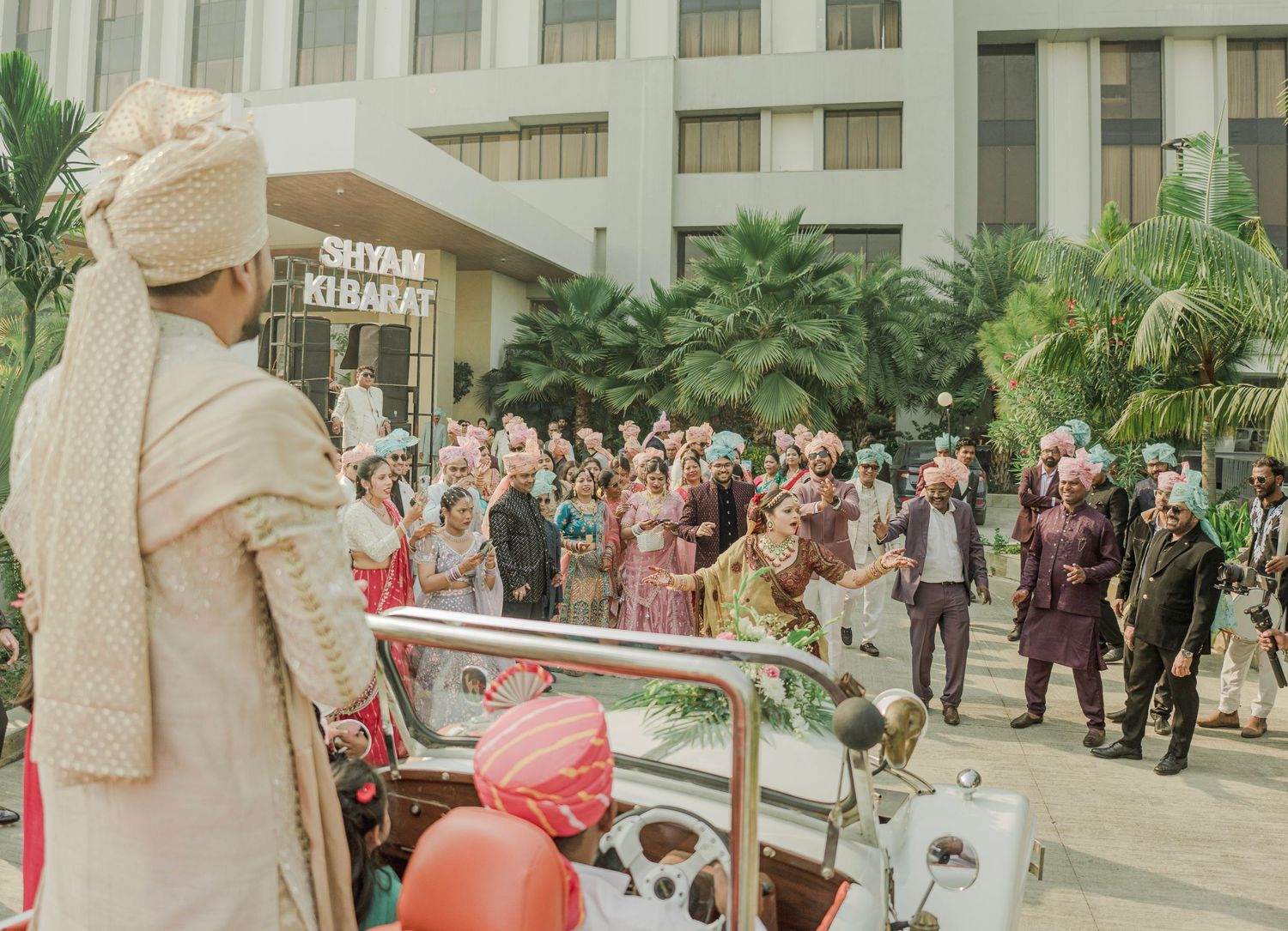 Groom in traditional turban watches baraat wedding procession with colorful guests outside hotel venue.