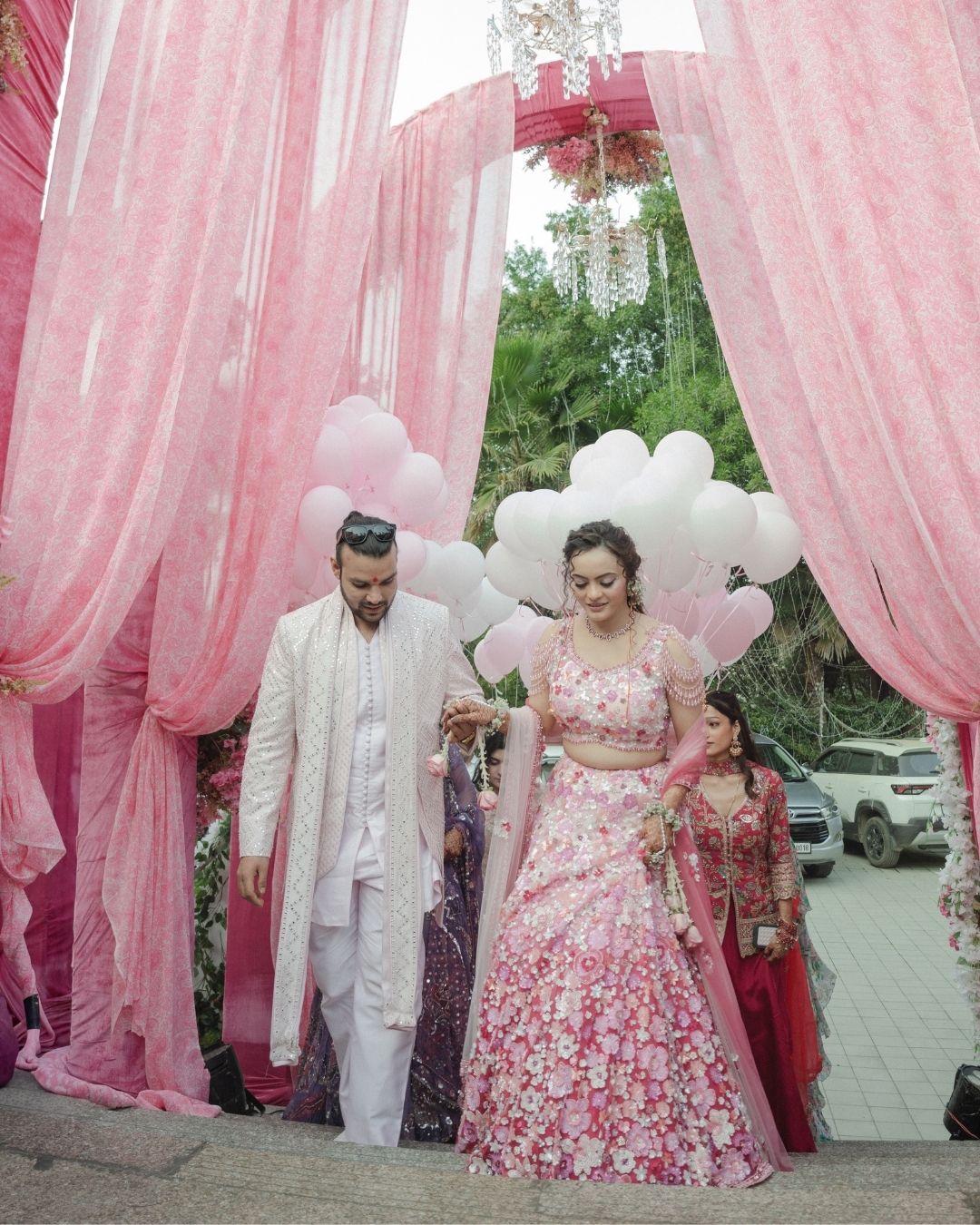Indian couple in traditional attire walking under pink drapes with white balloons at a wedding ceremony.