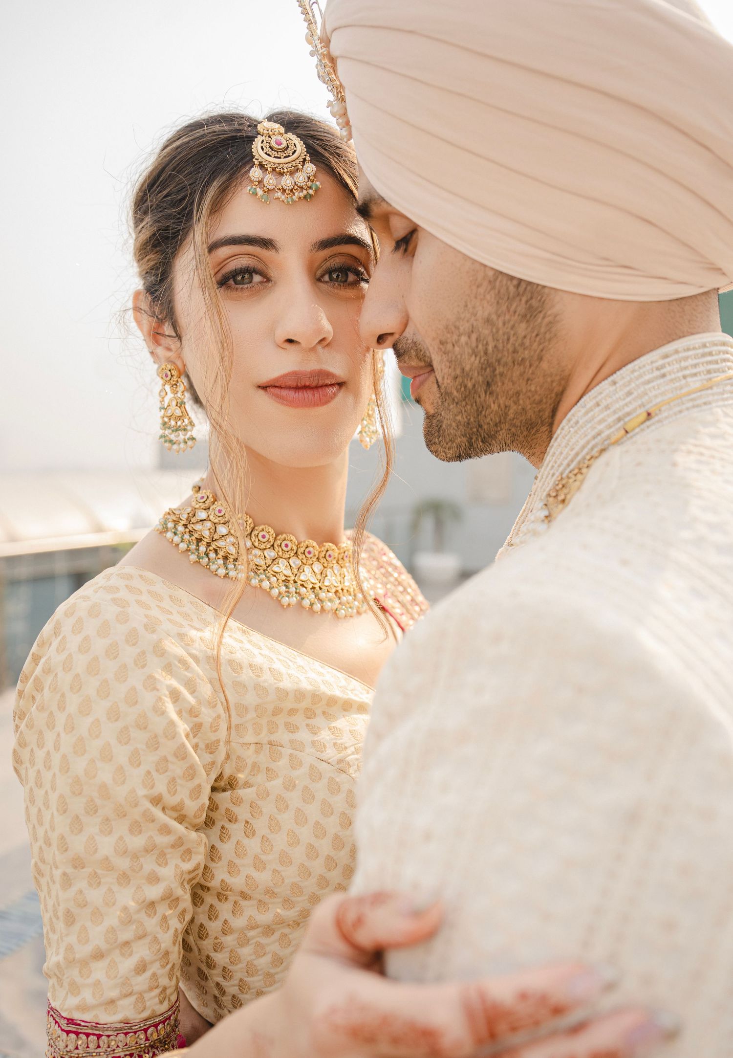 A Sikh bride in gold embroidered lehenga and kundan jewelry poses with groom in pink turban and sherwani.