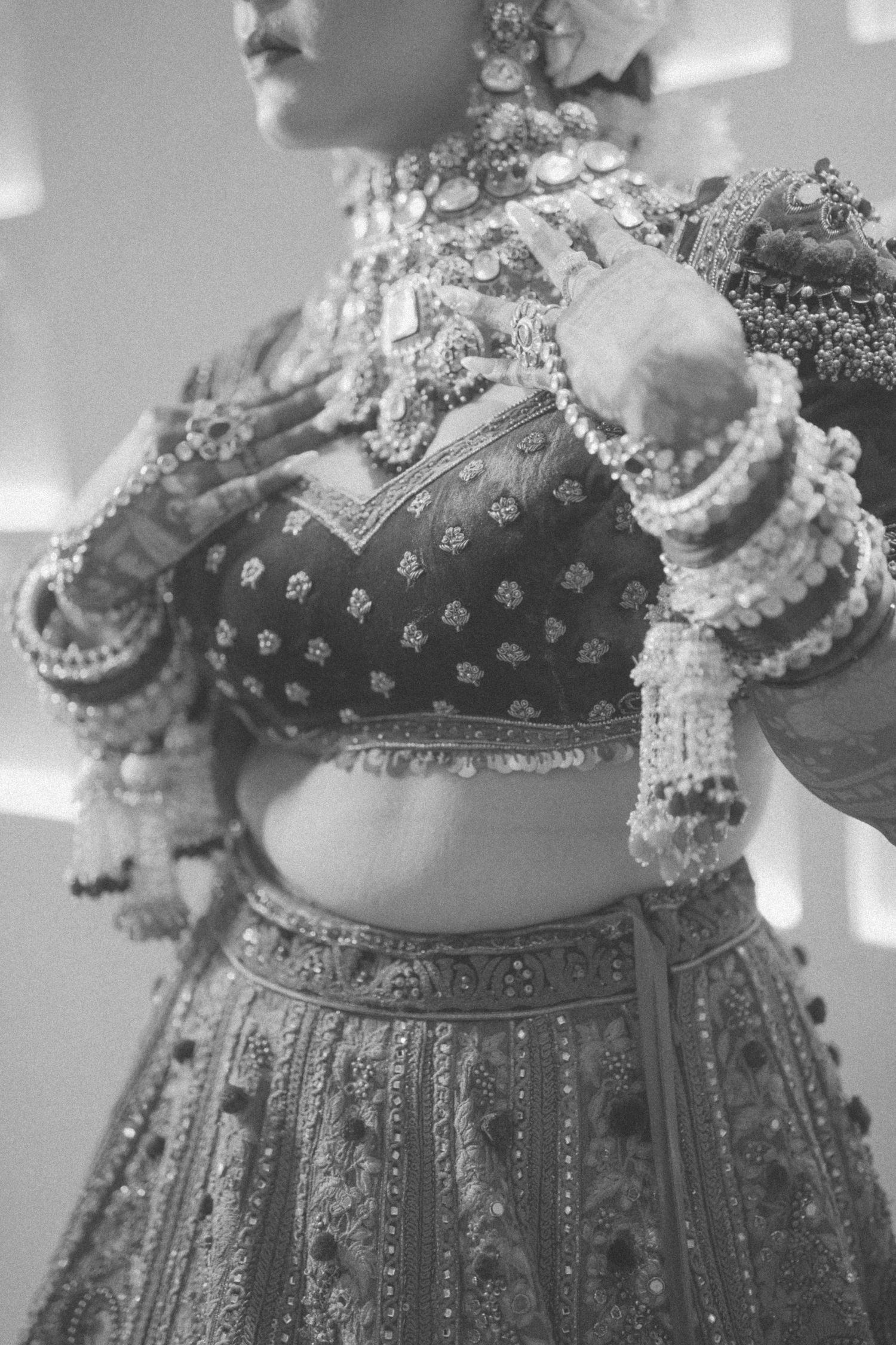 Black and white photo of a bride in ornate lehenga choli with heavy jewelry and bangles during Indian wedding.