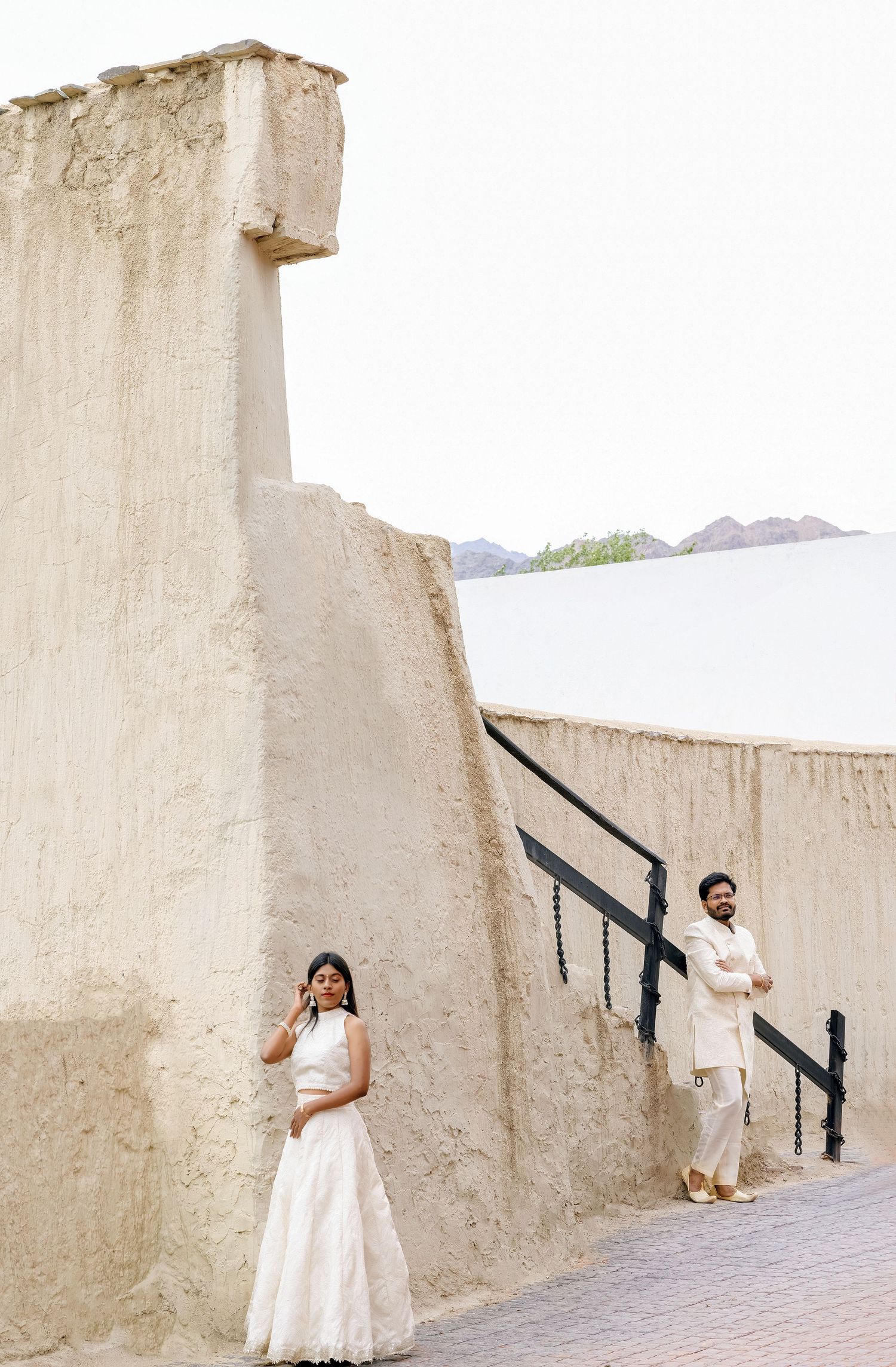 Couple in white attire posing by ancient beige stone walls with mountains in the background.