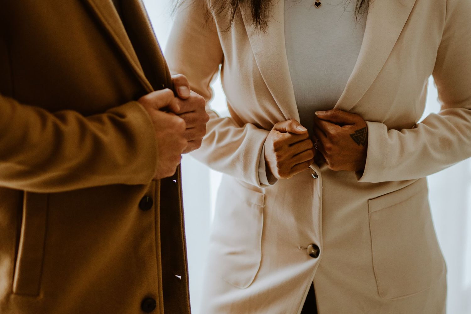 Two people in elegant beige suits adjusting jacket buttons, showcasing sophisticated formal fashion styling.