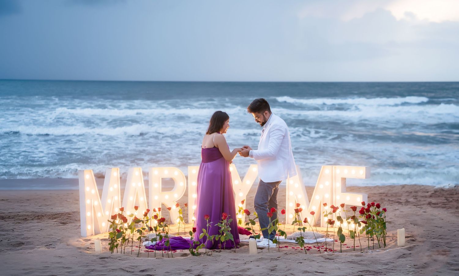 Romantic beach setup with illuminated 'MARRY ME' letters and floral decorations at sunset.