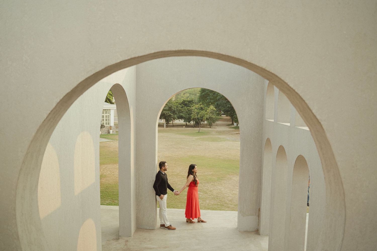Couple holding hands framed by white circular arches at an outdoor monument, woman in red dress, lush green lawn background.