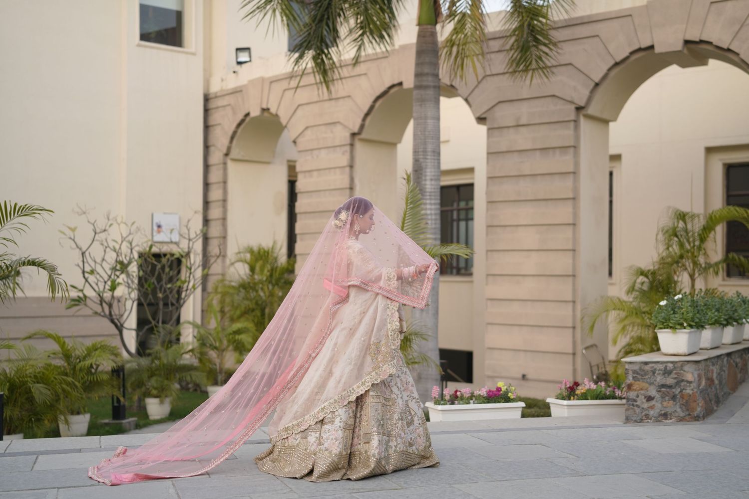 Bride in gold lehenga with flowing pink dupatta veil poses outdoors near elegant arched building.