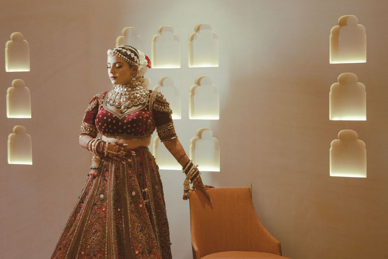 A bride in an ornate maroon lehenga and traditional jewelry poses elegantly in a warmly lit room with arched wall niches.