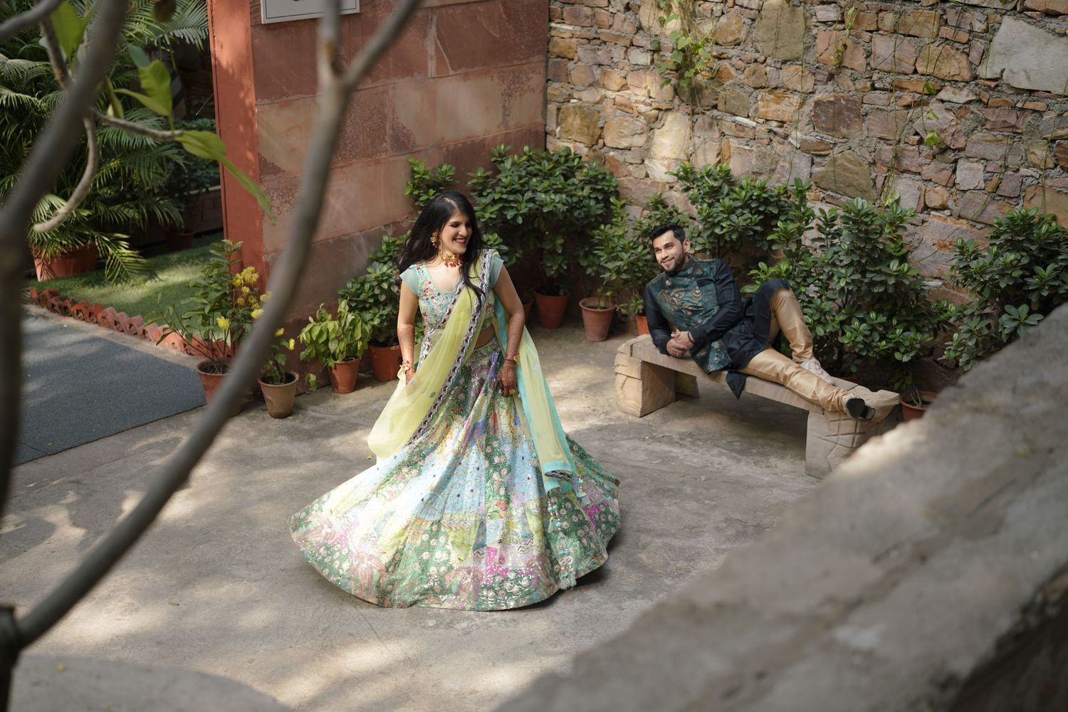 Bride in floral lehenga twirls as groom sits on bench in lush garden setting during Indian wedding photoshoot.