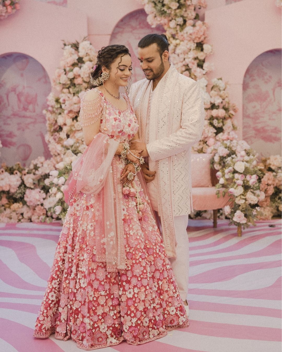 Indian couple in pink lehenga and white sherwani posing at floral wedding ceremony backdrop.