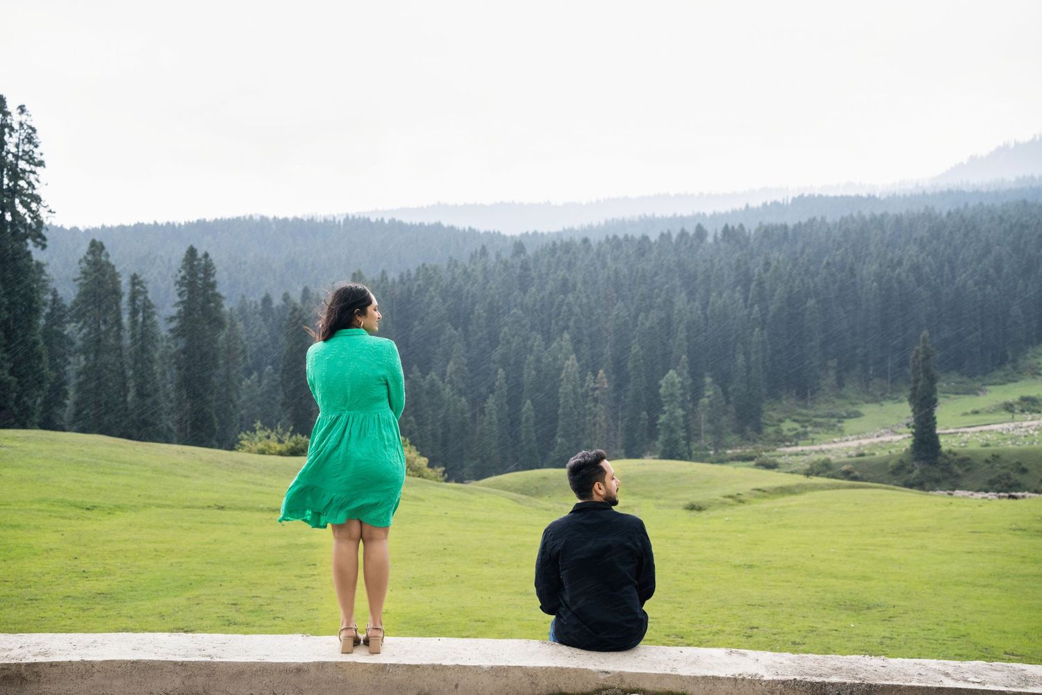 Couple in green dress and black outfit standing on stone ledge with pine forest and misty mountains backdrop.