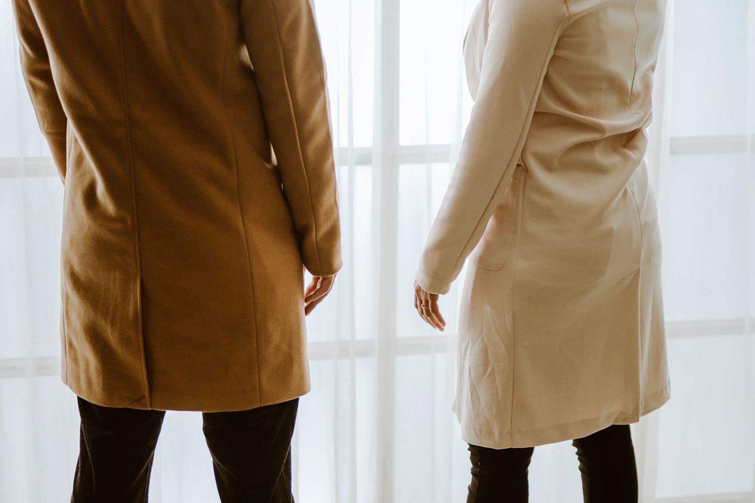 Two people standing side by side wearing elegant coats, viewed from behind near a bright window.