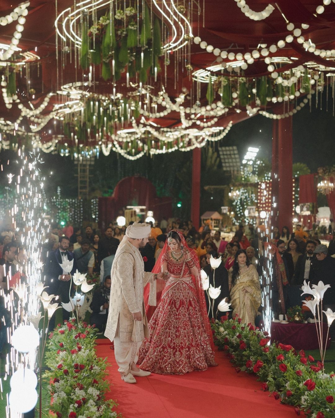 Bride in red lehenga and groom in sherwani walk through sparkling fireworks at an elaborately decorated Indian wedding.