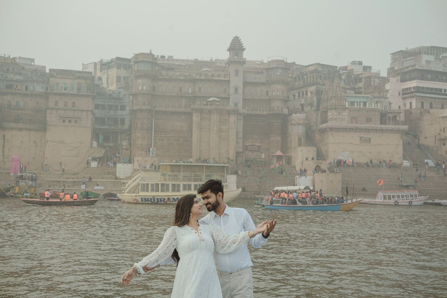 Couple in white attire posing romantically by the Ganges River with Varanasi ghats and boats in the misty background.