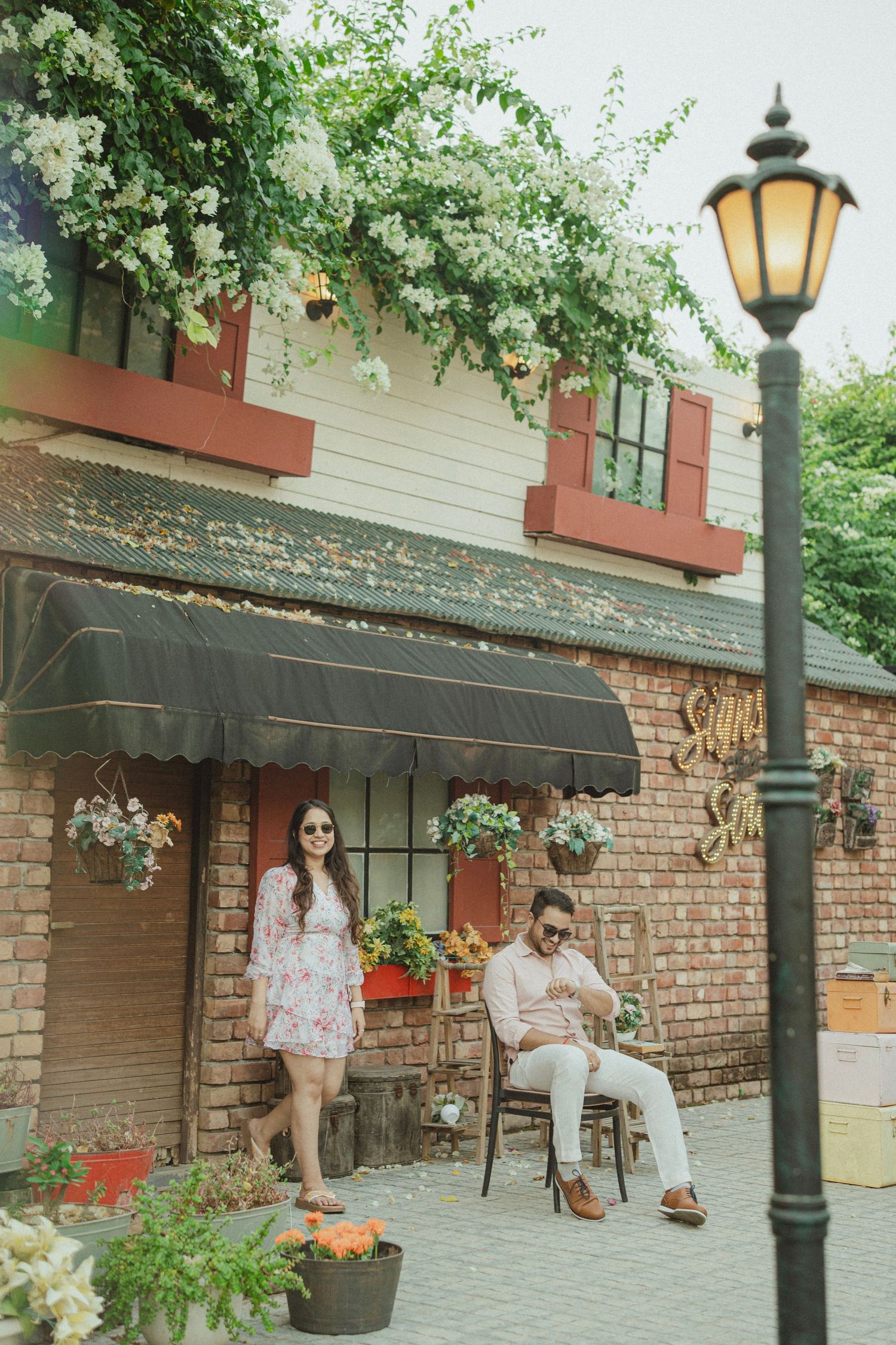 Couple posing outside a charming brick cafe with flower baskets, awning, and a vintage street lamp on a patio.