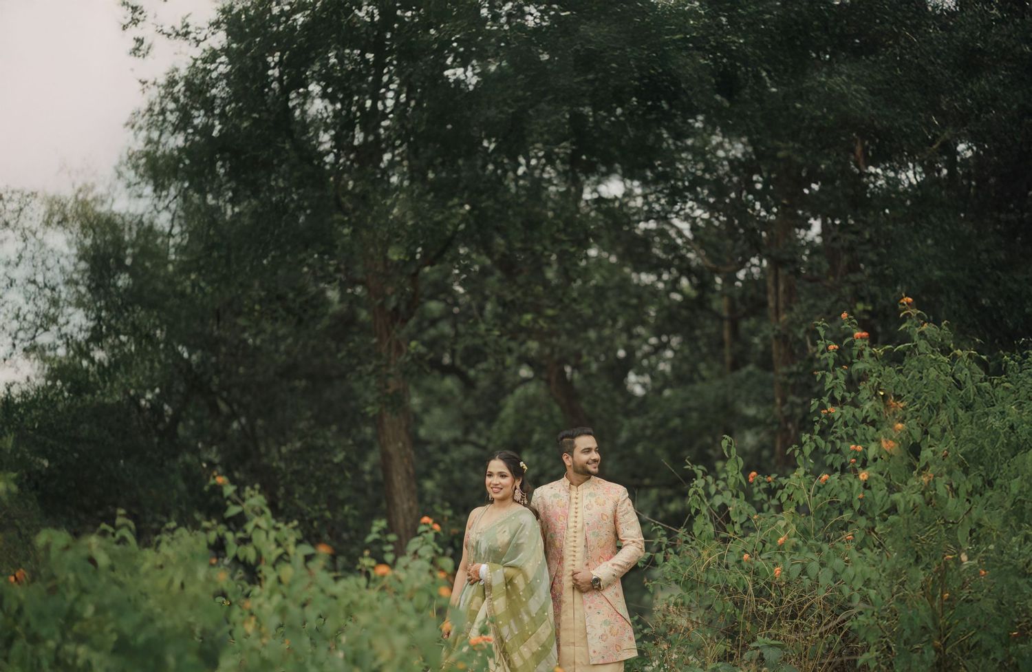 A couple stands together in a lush garden setting wearing traditional South Asian attire on an overcast day.