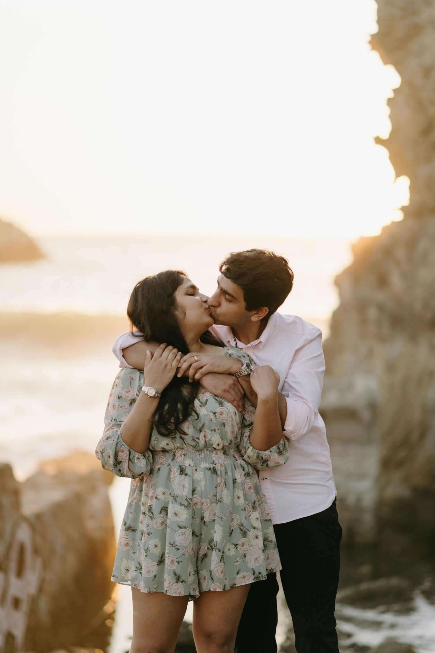 Couple embraces in soft sunset light against rocky coastal backdrop.