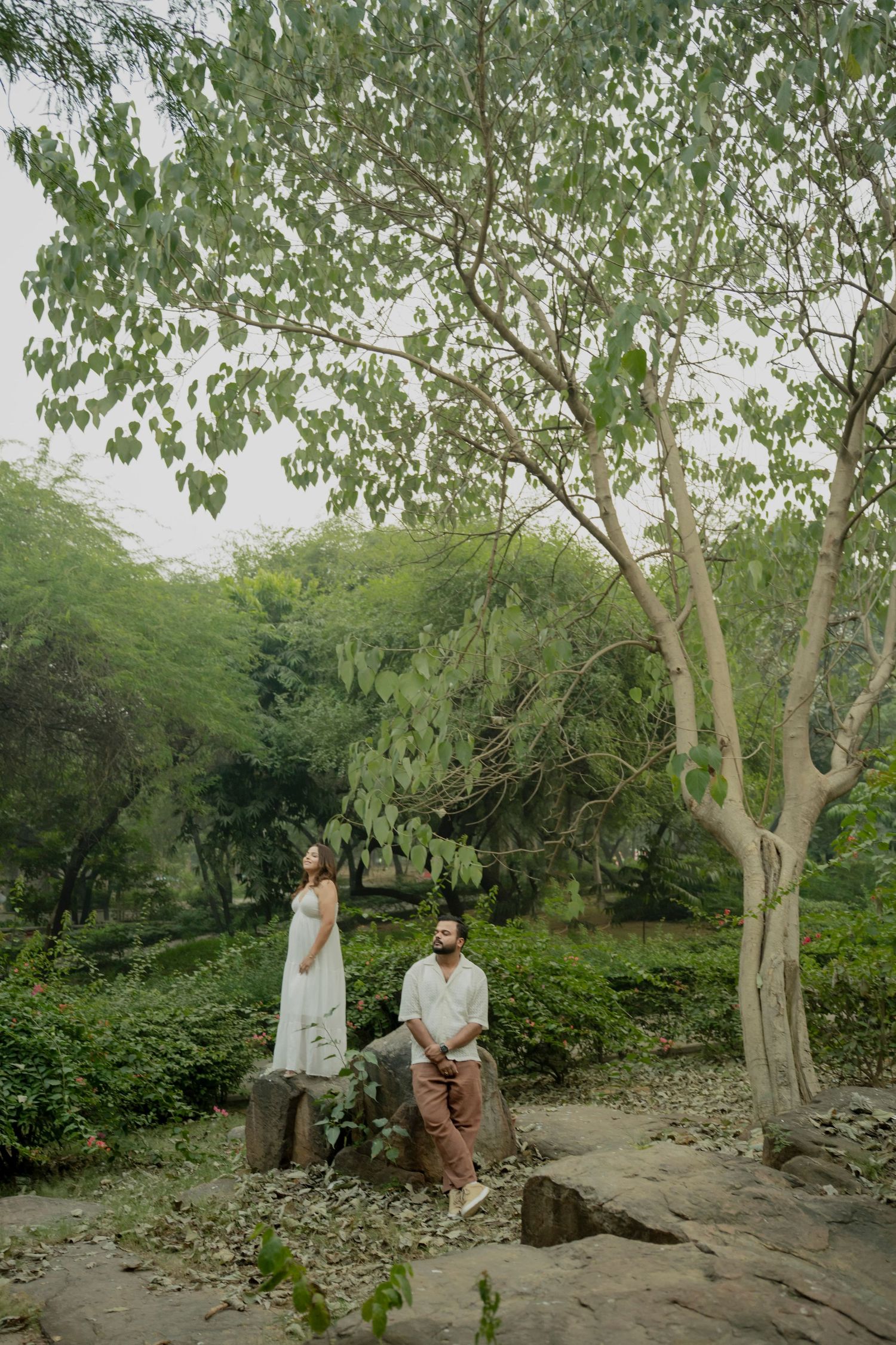 Couple in white outfits posing among lush green trees on rocky ground in a serene outdoor setting.