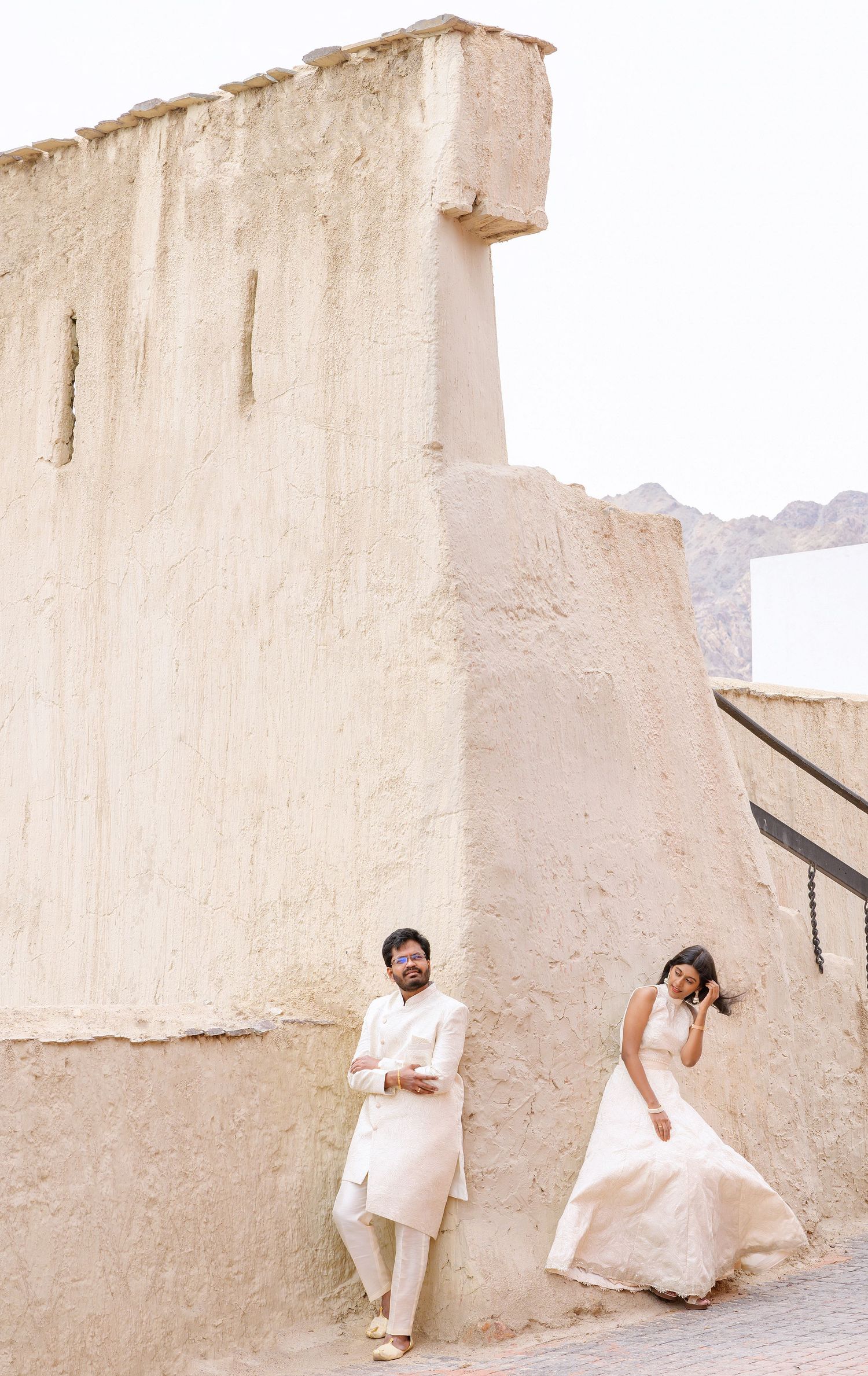 Couple in white wedding attire posing against ancient beige stone walls with mountains in the background.