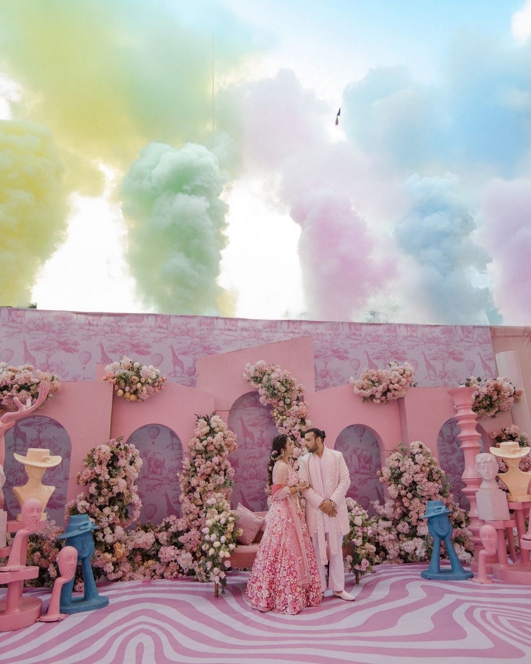 Couple in pink bridal lehenga and white sherwani pose before colorful smoke clouds at vibrant Indian wedding.