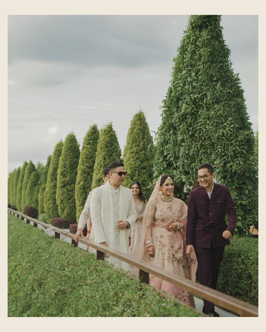 Three elegantly dressed guests walk along a manicured garden path lined with tall conical topiary trees at an Indian wedding.