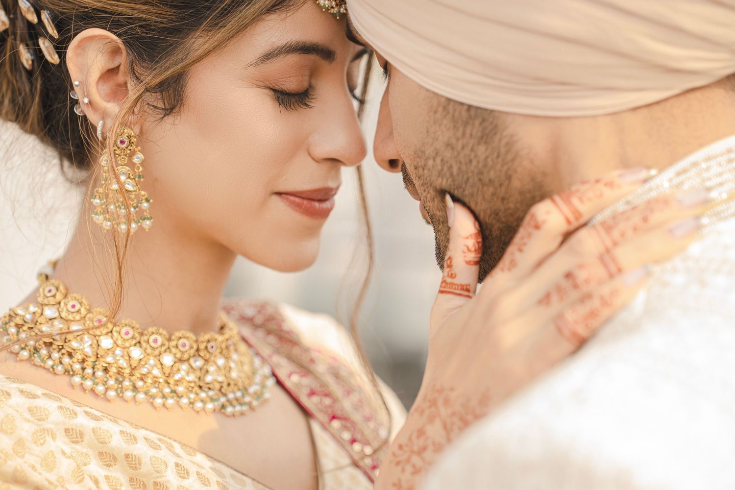 Indian bride and groom in traditional wedding attire share an intimate forehead touch, adorned with gold jewelry and mehndi.