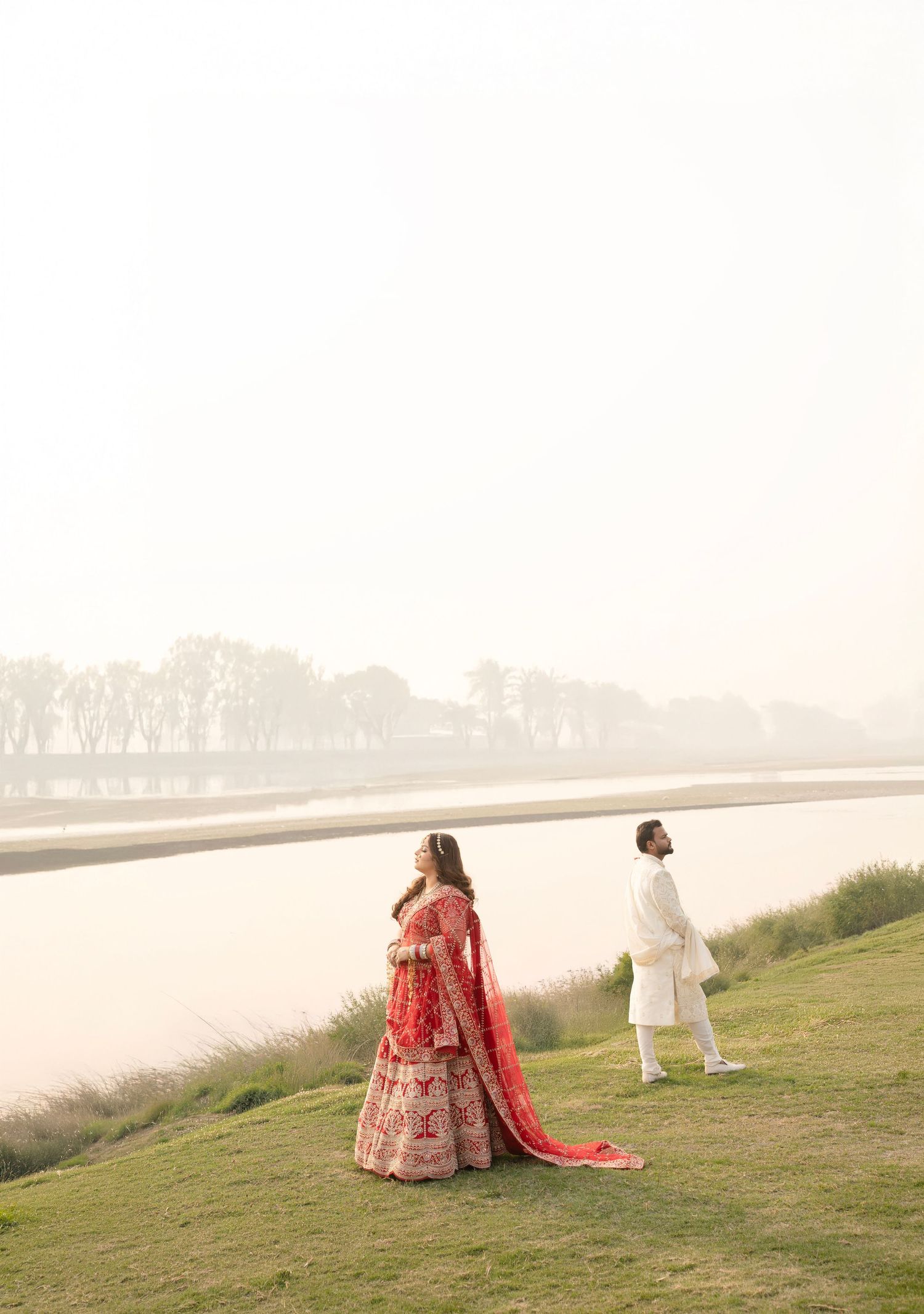 Indian bride in red lehenga and groom in white sherwani standing by a misty riverside during a traditional wedding shoot.