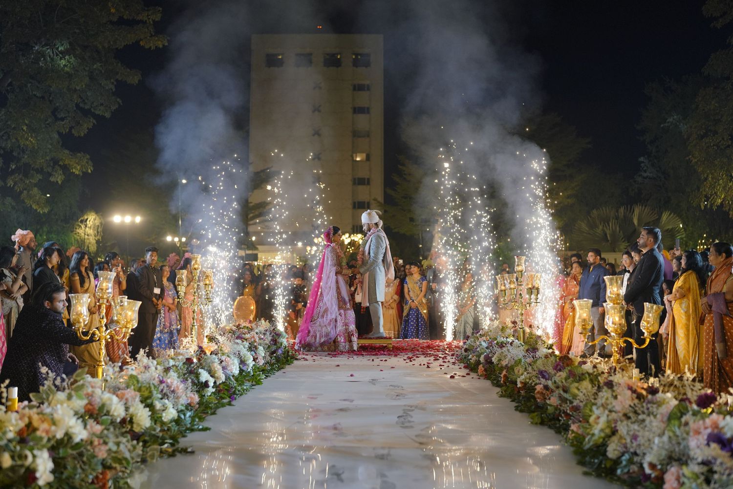 Bride and groom walk down floral aisle with sparkling fireworks at an elegant outdoor Indian wedding ceremony at night.