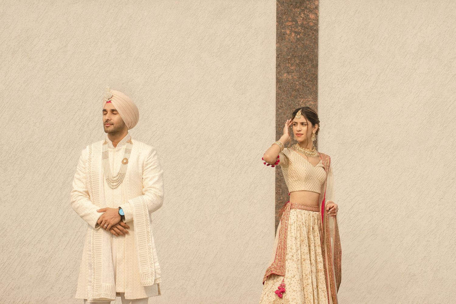 Indian couple in traditional wedding attire, groom in white sherwani and turban, bride in embroidered lehenga.
