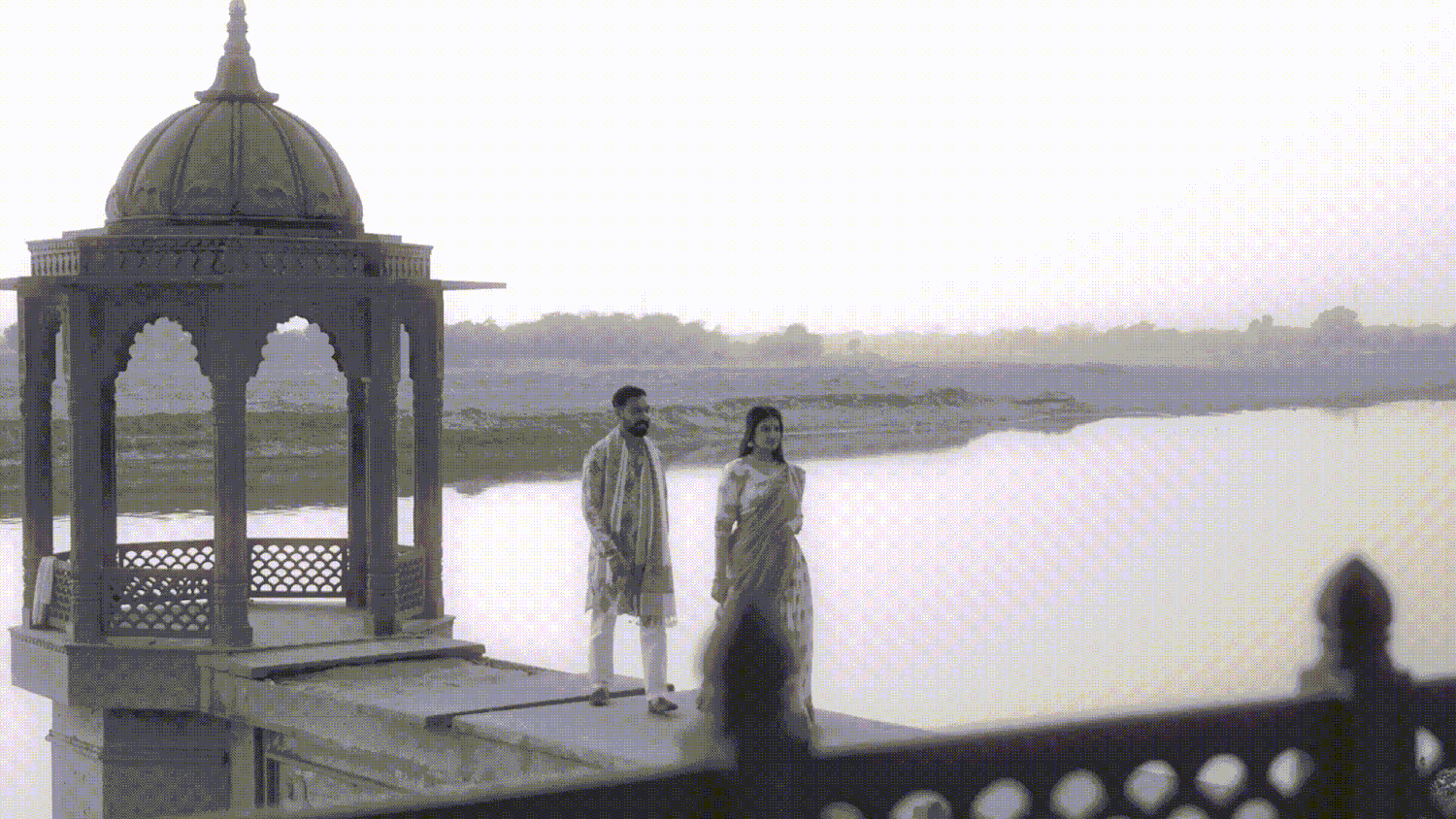 Two people stand under an ornate gazebo by a serene lake in a historic Indian architectural setting.