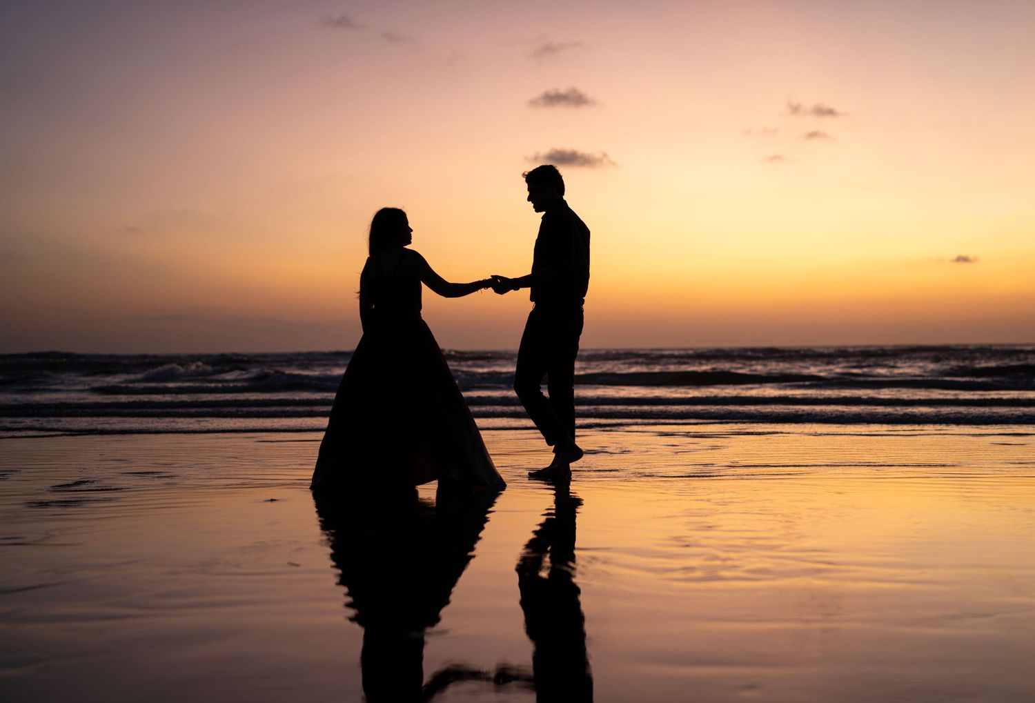Silhouettes against dramatic sunset sky on beach during proposal moment.